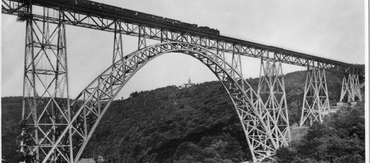 Müngstener Brücke mit Dampfzug, 1930–1960 Brücke aus Metall, die über ein Flusstal reicht. Auf der Brücke fährt ein Zug mit Dampflok