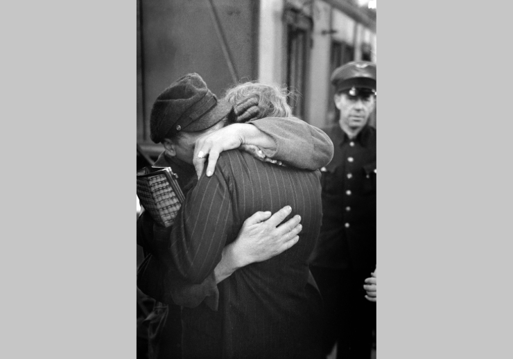 Mann und Frau umarmen sich an einem Bahnhof. Im Hintergrund ist ein Schaffner zu sehen.